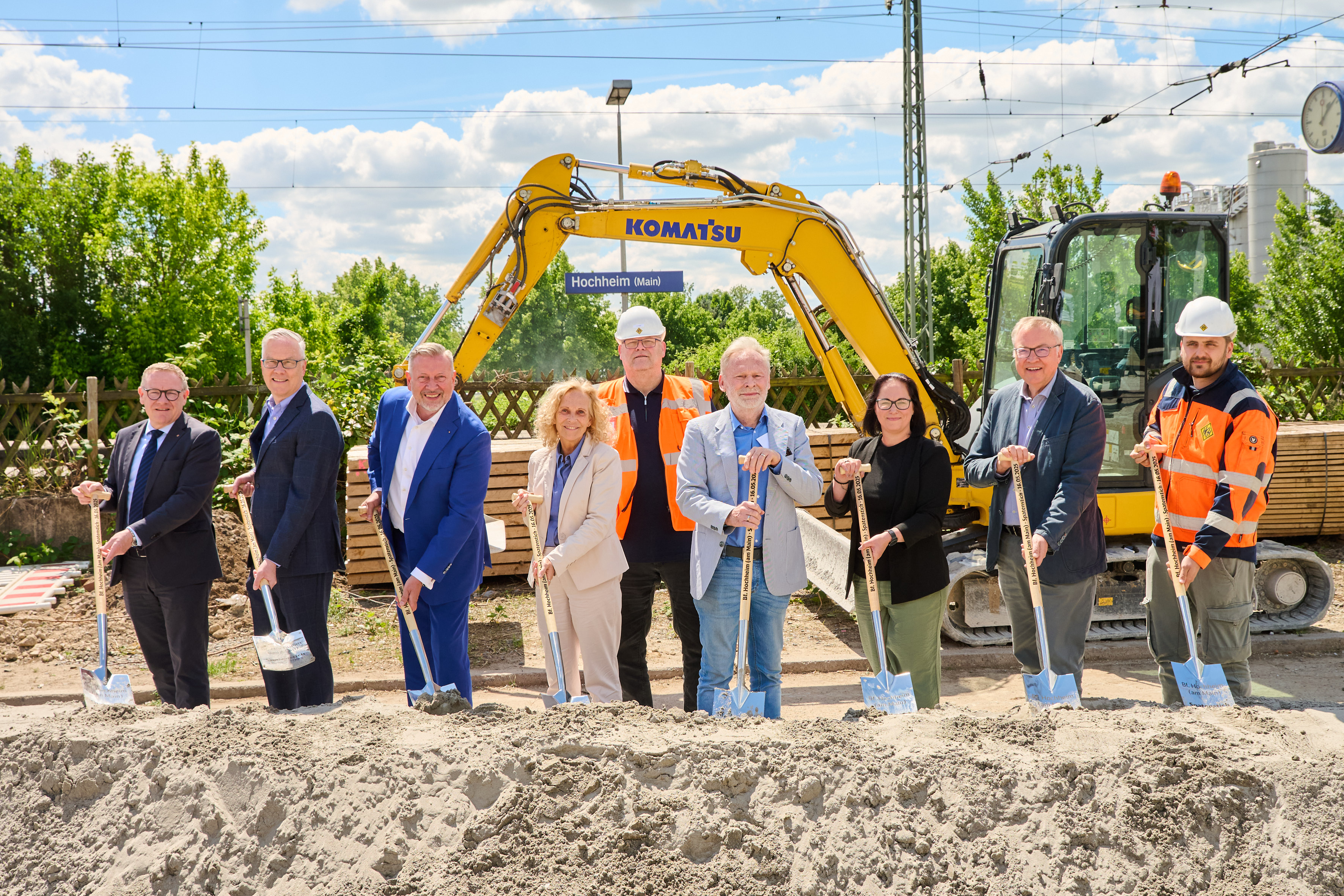 Neun Personen stehen nebeneinander stehen nebeneinander vor einem Sandhügel mit einer Schaufel in der Hand. Im Hintergrund das Stationsschild Hochheim