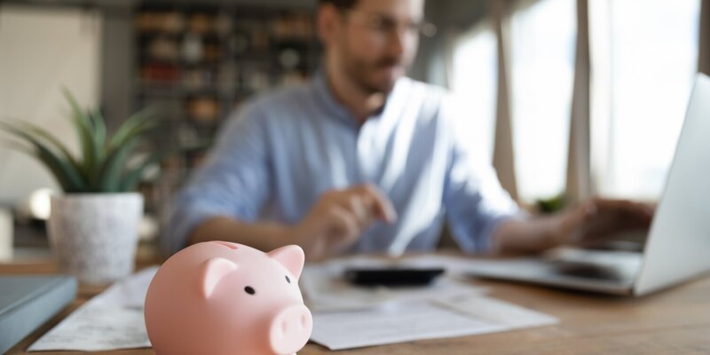 Sparschwein vor Mann mit Laptop, Taschenrechner und Unterlagen auf einem Schreibtisch