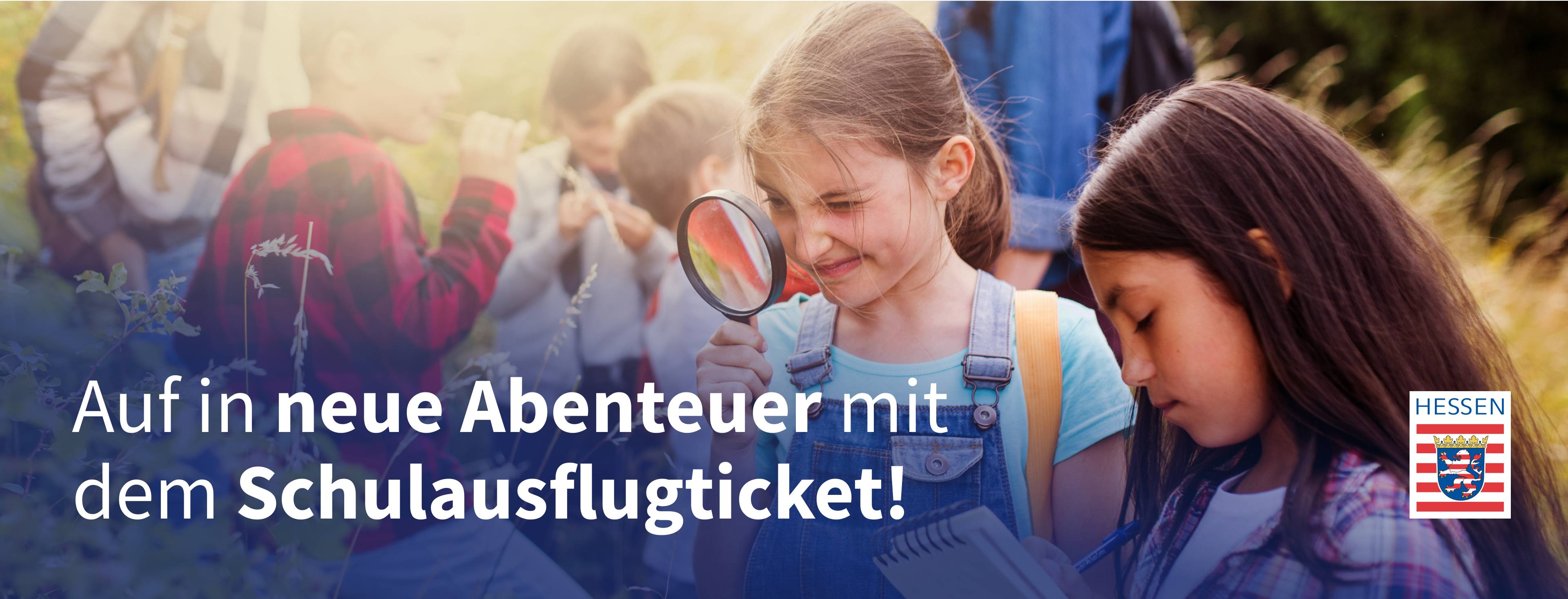 Zwei Mädchen mit Lupe und Notizblock betrachten draußen Blumen, im Hintergrund sind weitere Kinder und zwei Aufsichtspersonen.